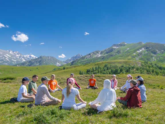 Группа людей сидит в кругу и медитирует на зеленой поляне на фоне гор