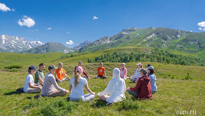 Группа людей сидит в кругу и медитирует на зеленой поляне на фоне гор 